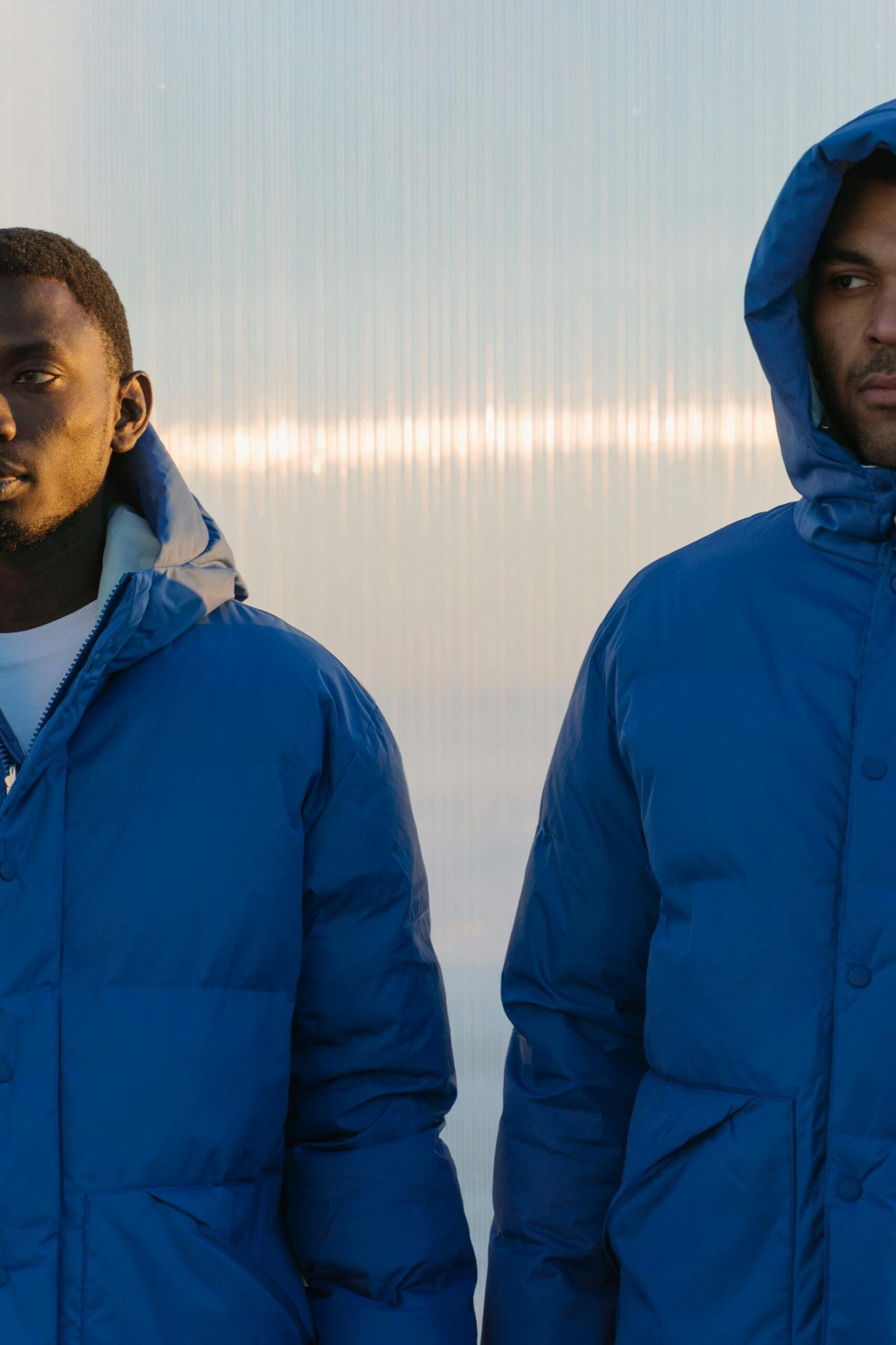 Two men in blue puffer jackets stand against a glass wall creating a modern aesthetic.